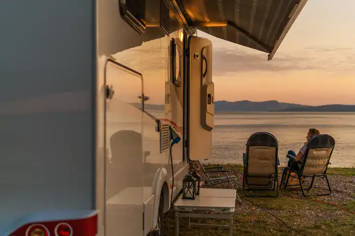 Fifth wheel parked up by lake with people sitting in chairs outside watching the sunset.