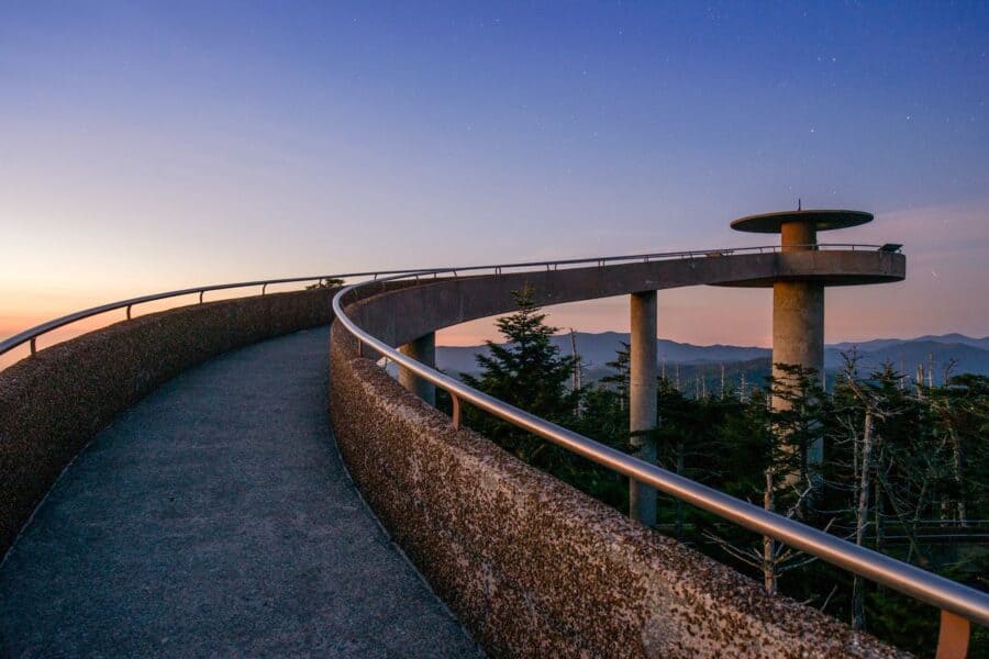 Twilight view from Kuwohi, formerly Clingmans Dome observation tower, featuring a curved walkway with textured railing leading towards an elevated round platform, set against a backdrop of a gently fading sky and distant mountain silhouettes.