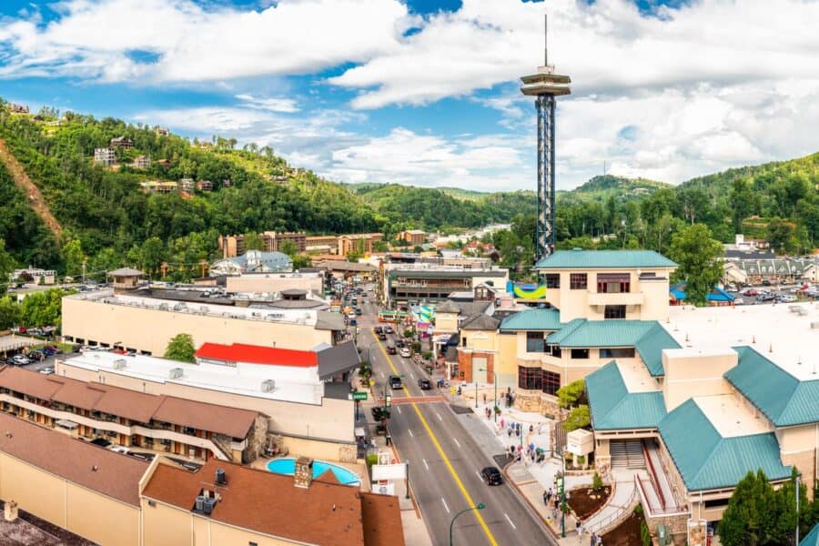 Aerial view of downtown Gatlinburg, Tennessee, showcasing a bustling main street with colorful buildings, various shops, and attractions, including the distinctive Gatlinburg Space Needle. The town is nestled in a valley surrounded by lush green hills, illustrating a vibrant blend of urban activity and natural scenic beauty.