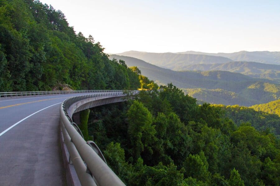 Scenic view from Foothills Parkway near Gatlinburg, Tennessee, showing a curving road hugging the side of a lush, green mountain. The road overlooks expansive layers of forested hills stretching into the distance, bathed in soft sunlight, offering a stunning vista of the Smoky Mountains.