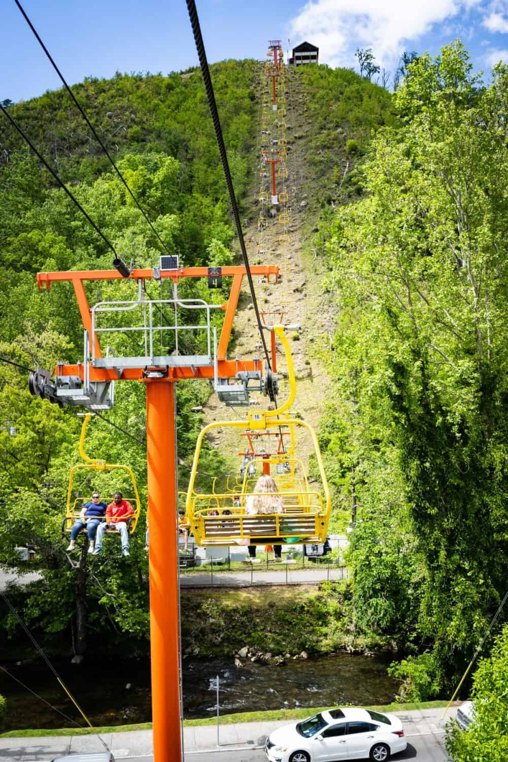 View from the Gatlinburg Skylift showing brightly colored yellow chairlifts ascending a lush green mountain. Passengers enjoy the ride overlooking a small creek and a street below, with the hilltop station visible in the distance against a clear blue sky.