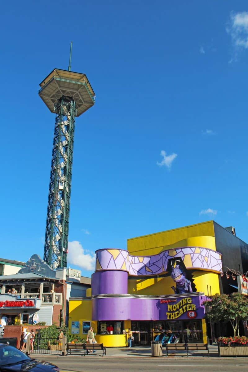 The Gatlinburg Space Needle, a tall observation tower, rises above a colorful urban setting under a clear blue sky. Below, the vibrant yellow and purple facade of a building advertises a 5D Moving Theater, bustling with people and activity, capturing a lively street scene in downtown Gatlinburg, Tennessee.