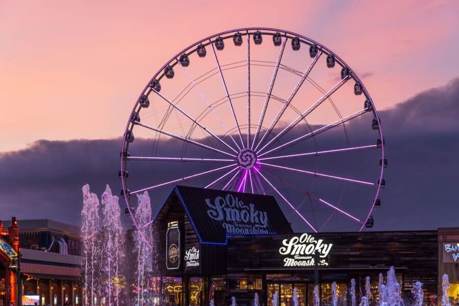 Evening view of the Great Smoky Mountain Wheel in Pigeon Forge, illuminated with vibrant purple lights against a dramatic sunset sky with pink and purple hues. The large Ferris wheel dominates the scene, with the Ole Smoky Moonshine building and playful water fountains in the foreground, creating a lively and picturesque atmosphere.