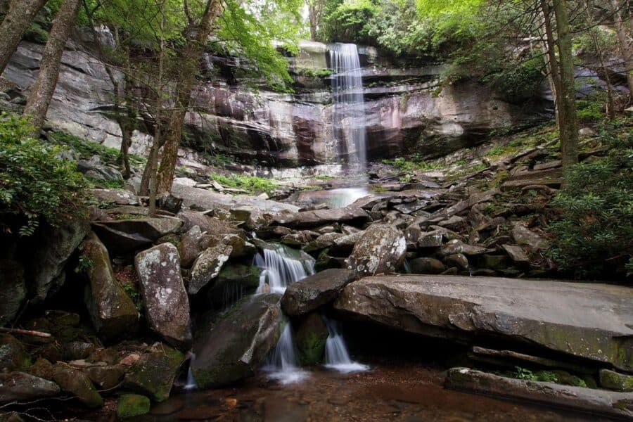 Rainbow Falls in the Great Smoky Mountains National Park, showcasing a tranquil waterfall cascading down a large rock face surrounded by lush greenery. The waterfall creates a misty veil over a pool below, with large, jagged rocks scattered around the area, contributing to the serene and natural beauty of the scene.