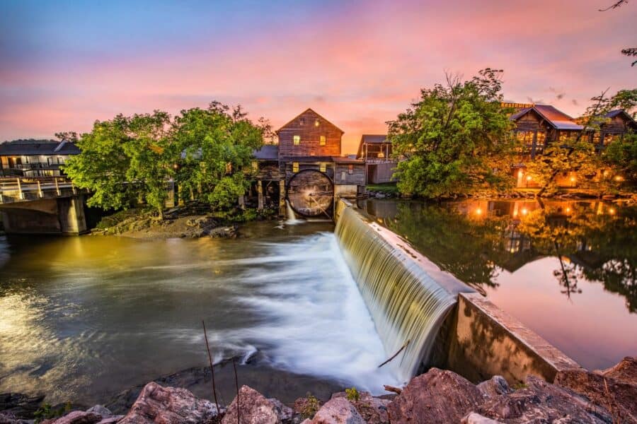 Picturesque view of an old mill with a functioning water wheel next to a cascading waterfall, captured during sunset in Gatlinburg, Tennessee. The scene includes a rustic building illuminated by warm lights, reflecting beautifully on the calm water surface against a vibrant pink and blue sky.